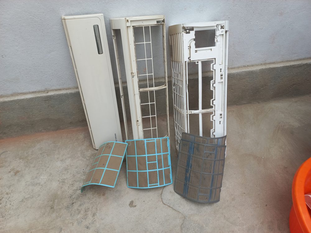 Tampa, grelha e filtros de uma unidade interior desmontada, encostados à parede para limpeza